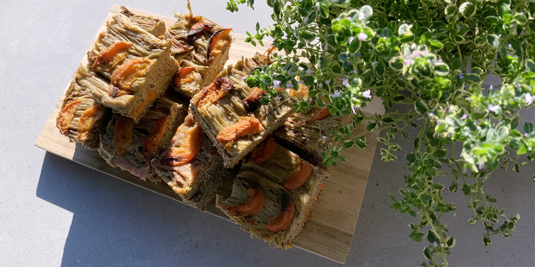 Stücke von einem saftigen Aprikosen-Mohn-Rhabarber-Kuchen unter einem Strauß Zweige & Blumen auf einem Holzbrett in der Sonne.