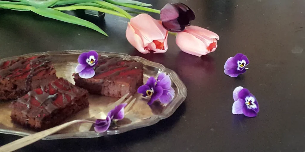 Ein silberner Teller mit drei Stücken Rote Beete Brownies und einer Gabel, gerahmt von violetten Stiefmütterchen vor drei rosa-lila-farbenen Tulpen auf einem Tisch.